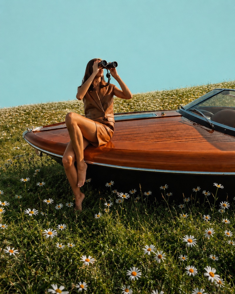 A woman in a leather set looks through binoculars while sitting on the bow of a wooden speedboat in a daisy field